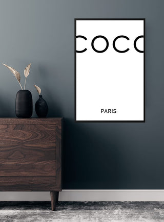 White COCO poster with minimalist text "COCO PARIS" hanging on a dark wall above a wooden dresser.