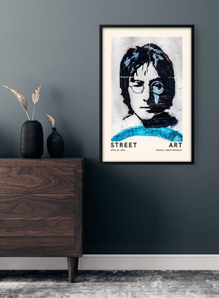 Framed John Lennon street art poster hanging on a dark wall above a wooden dresser with decorative vase.