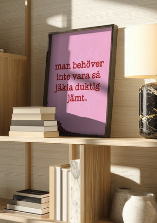 Duktig jämt by Grafik bageri in a dark frame on a wooden shelf beside books and a lamp