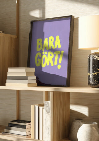 Bara gört! by Grafik bageri in a dark frame on a wooden shelf beside books and a lamp