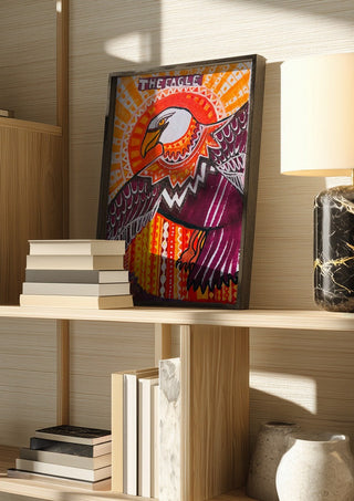 Eagle in the sky by ALEX in a dark frame on a wooden shelf beside books and a lamp