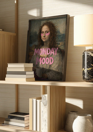 Monday Mood by Decker Studios in a dark frame on a wooden shelf beside books and a lamp