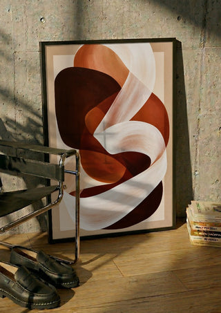 Ribbon in Rust by Martina Karlsson in a black frame leaning against a concrete wall by a steel chair
