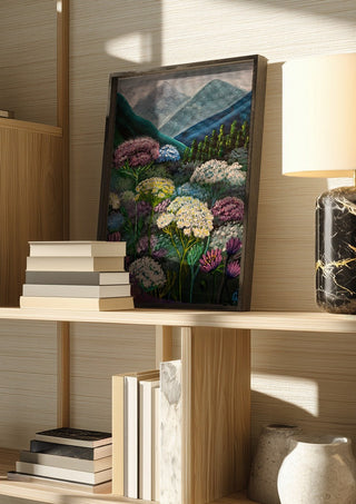 Mountain Meadow by Svetla Stoyanova Bassi in a dark frame on a wooden shelf beside books and a lamp