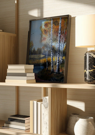 Autumn Birches by the Pond by Lüba in a dark frame on a wooden shelf beside books and a lamp