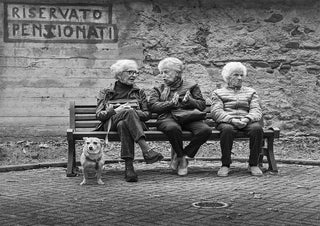 Three elderly women sit on a bench with a dog, under a sign reading "Riservato Pensionati" (Reserved for Retirees).