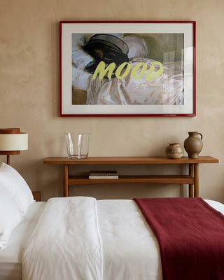 Framed "Woman In Repose" by Talitha McQueen above a bed with white linens and a burgundy throw.