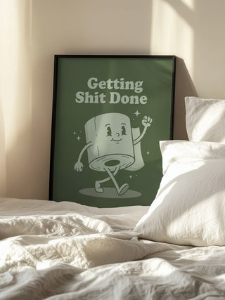Framed "Getting Shit Done" poster with cartoon toilet paper roll on display by a white bed.