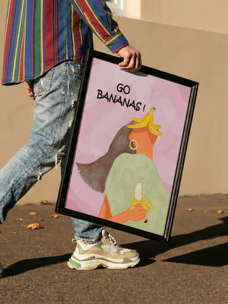 Person holding framed "Go Bananas Playful" art print by Sannel Larson featuring a woman with bananas on her head.