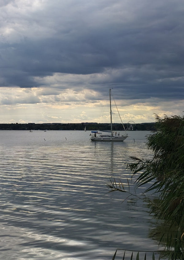 Light Reaching the Water by Bora Kőhalmi - a white sailboat under a heavy grey cloud with a band of light over Lake Balaton