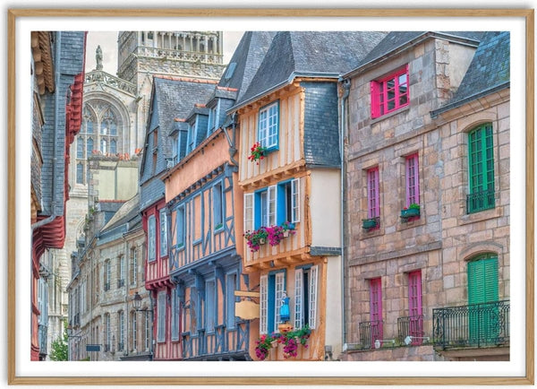 Colorful historic buildings line a quaint street in an old French town, with flowers and cathedral in the background.